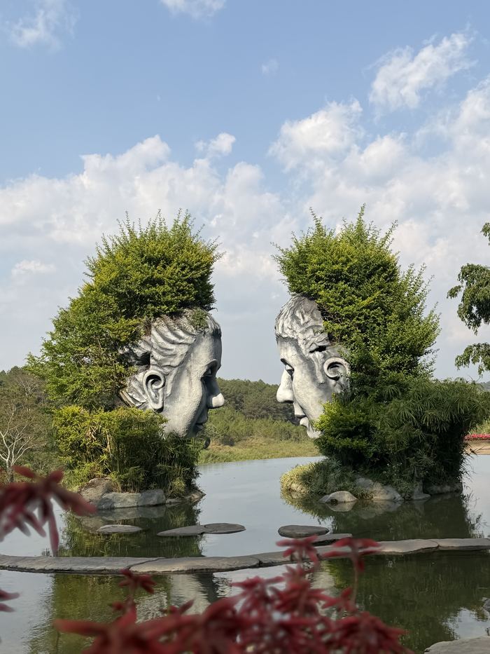 Two face sculptures with plant hair facing each other across a pond